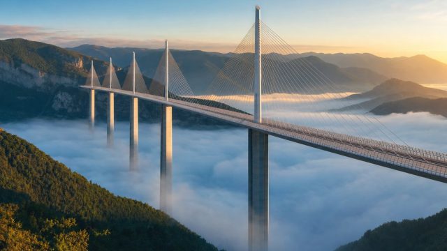 Viaduct modern cu piloni înalți traversând o vale acoperită de ceață la răsărit